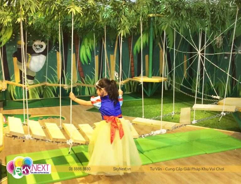 Bé gái mặc váy công chúa bám dây đi trên cầu gỗ treo trong khu vui chơi
