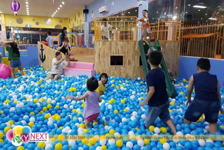 Trẻ em chơi trong bể bóng xanh, trắng, vàng cạnh cầu trượt và khung gỗ