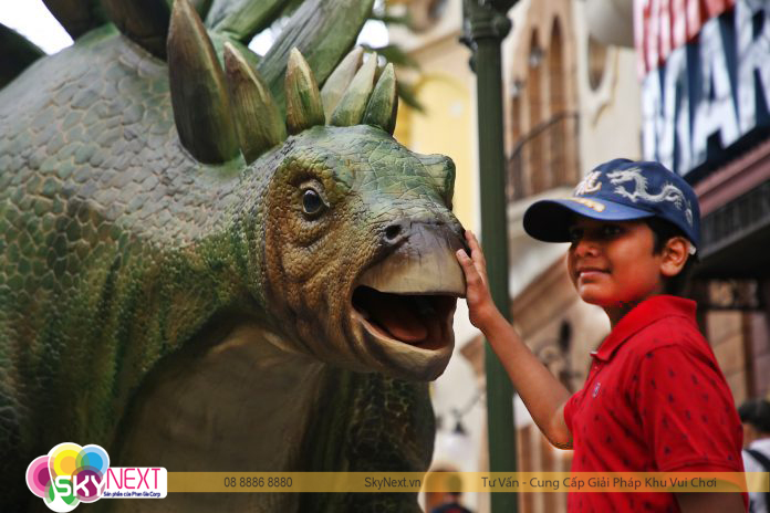 Trẻ em chạm tay vào mô hình khủng long Stegosaurus xanh trong khu vui chơi