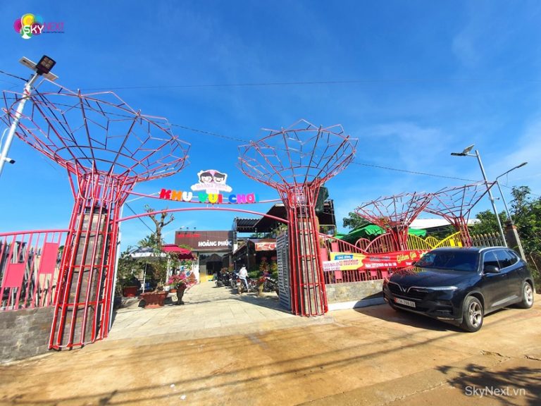 Cổng vào khu vui chơi với khung kim loại đỏ hình cây và ôtô đỗ trước
