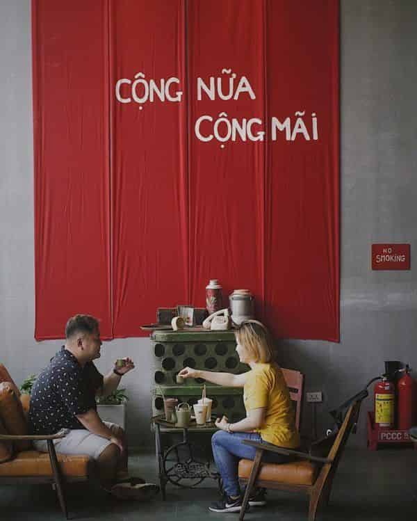 Hai khách ngồi trước tấm rèm đỏ có dòng chữ 'CỘNG NỬA CỘNG MÃI'