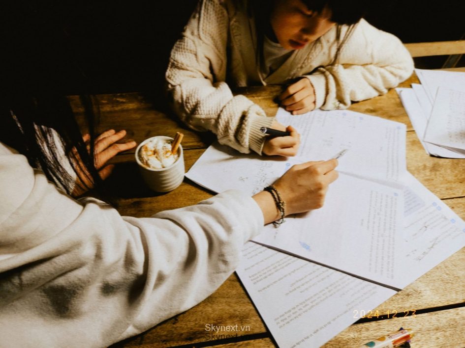 Hai người ngồi bên bàn gỗ, viết ghi chú trên giấy cạnh tách cà phê