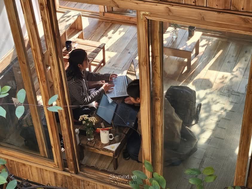 Hai người ngồi trên sàn gỗ trong quán, làm việc với laptop bên cửa kính